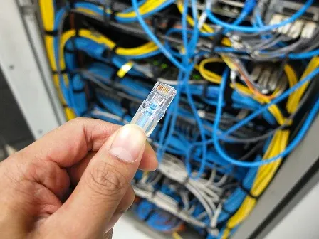 Hand holding an Ethernet (RJ45) connector in front of a network cabinet filled with blue and yellow cables